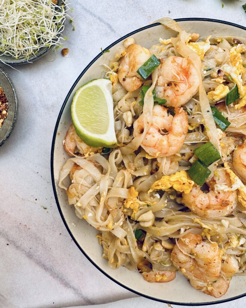 a close up on a plate with pad thai, rice noodles with prawns, egg, lime and pad thai sauce